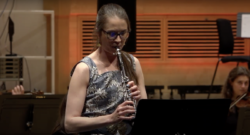woman with glasses and hair tied back playing clarinet, looking concentrated. Photo is from waist-up