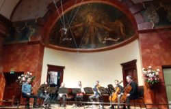 6 musicians sat in semicircle onstage at wigmore hall. White background wall with beautiful art=covered half-dome ceiling